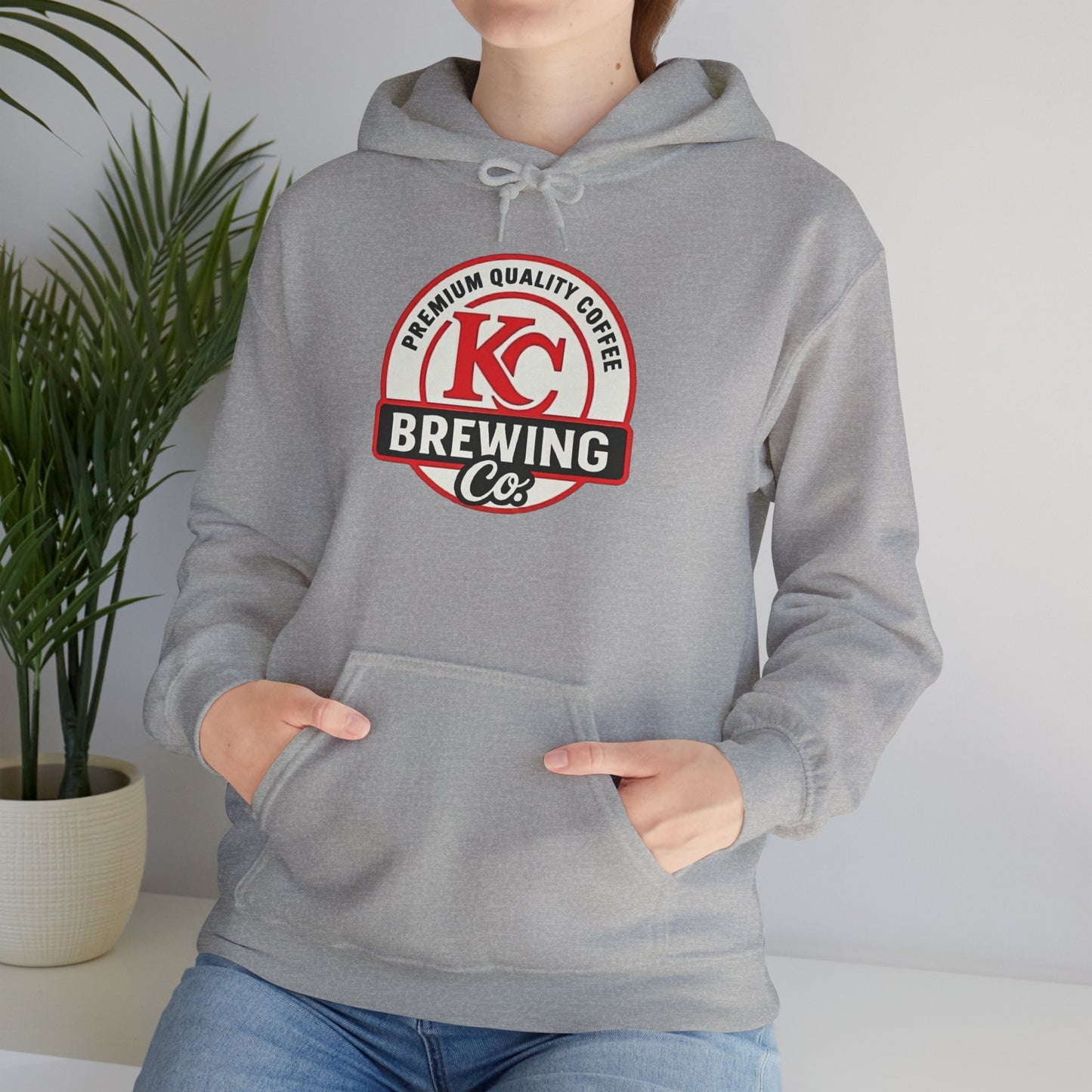 Person wearing grey KC Brewing Co hoodie with premium quality coffee logo indoor near potted plant