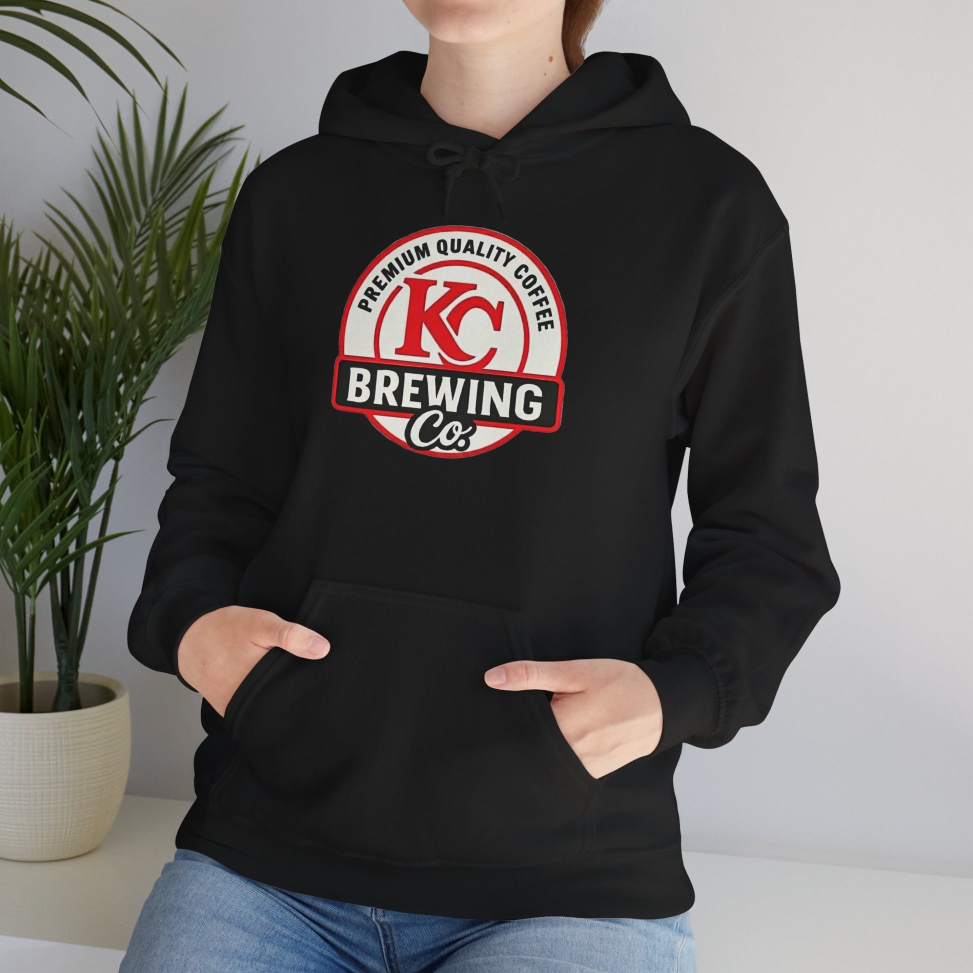 Person wearing black hoodie with KC Brewing Co premium quality coffee logo sitting indoors next to green plant