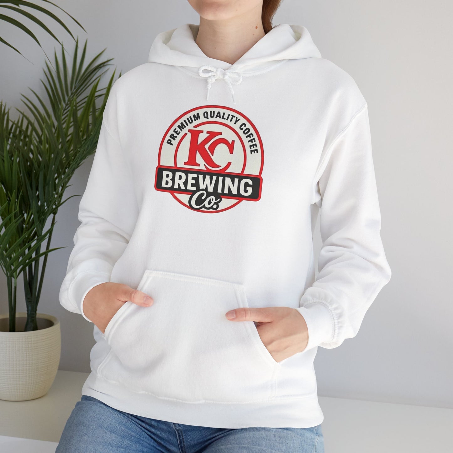 Person wearing white hoodie with KC Brewing Co premium quality coffee logo, indoor plant in background